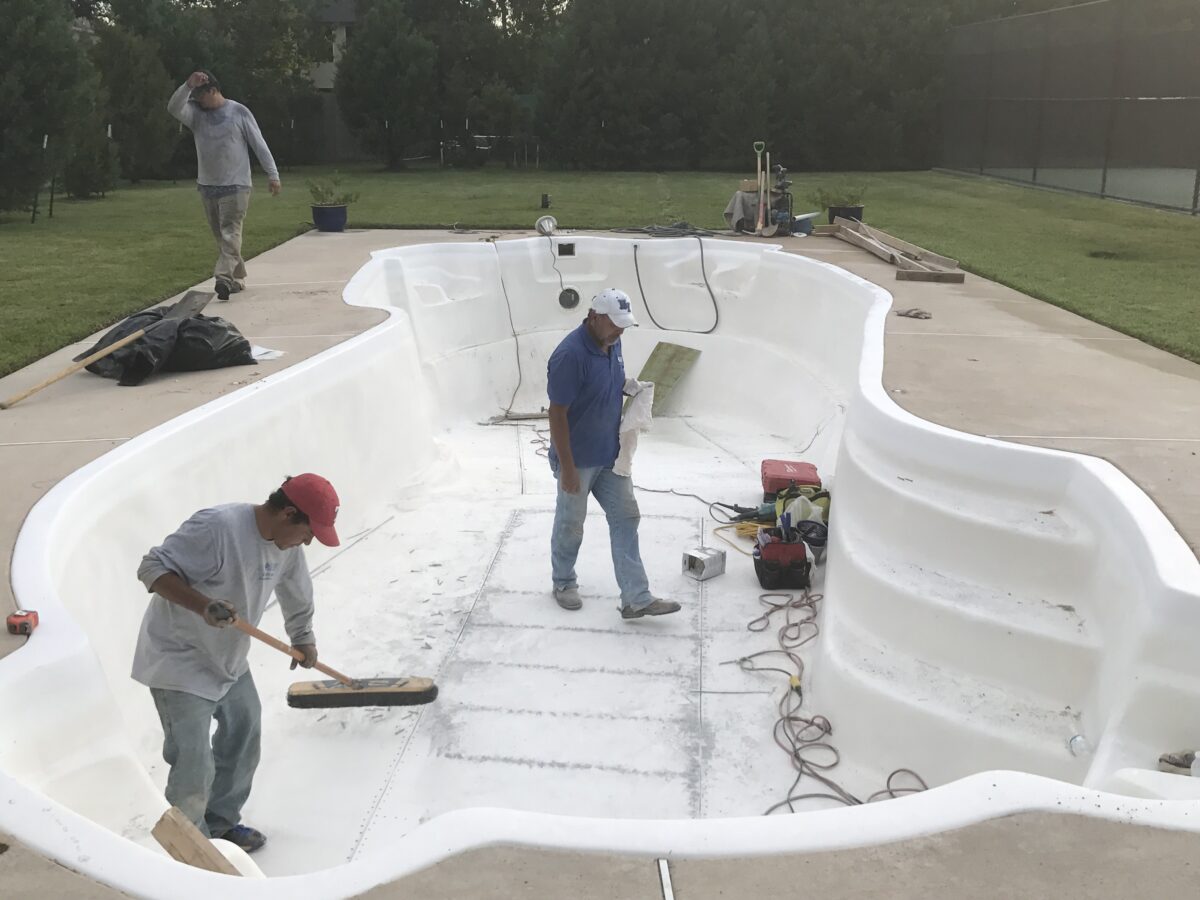 Workers renovating an in ground swimming pool in a backyard. preparing it for the next step in the process. the background deck is tan surrounded by a grass lawn