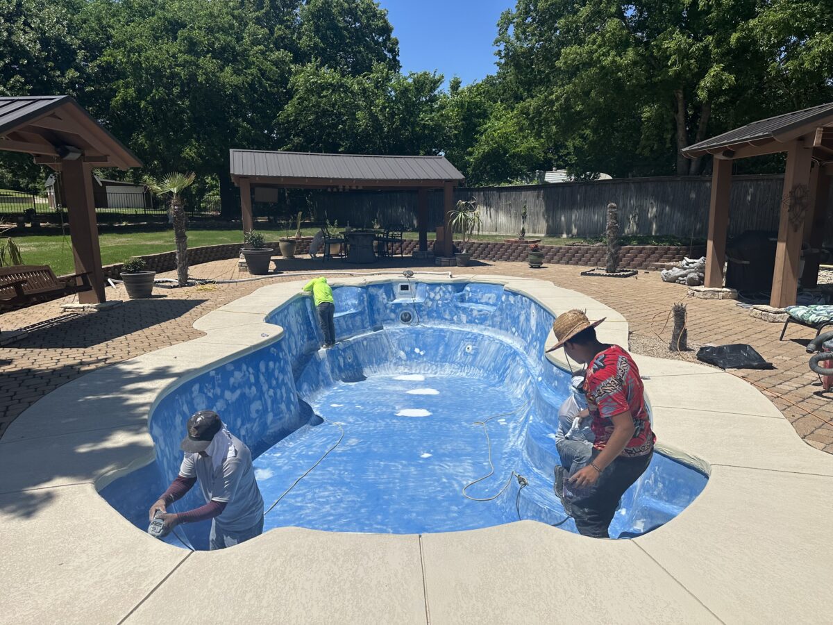 Workers renovating an in-ground swimming pool in a backyard, with the pool surface being prepared for a new finish and pool repair. The surrounding area features a paved deck, outdoor seating, and a pergola with a covered patio.